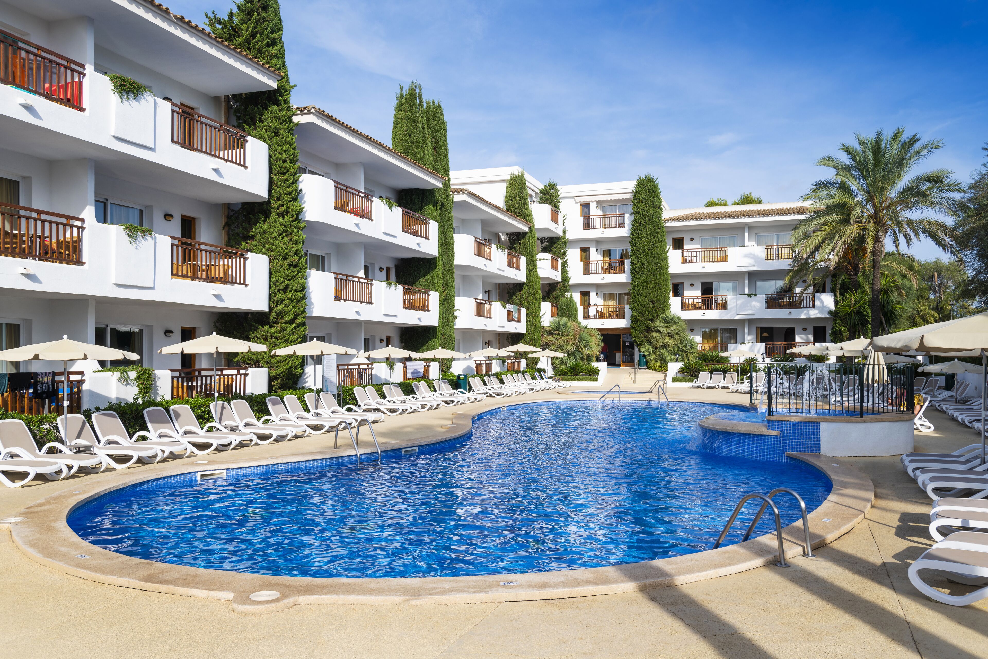 A swimming pool surrounded by white lounge chairs and umbrellas, with multi-story buildings in the background.