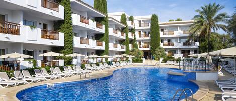 A swimming pool surrounded by white lounge chairs and umbrellas, with multi-story buildings in the background.