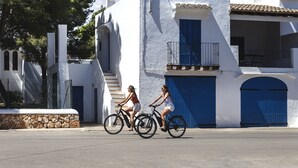 Two women riding bicycles on a street with white buildings and blue doors. - Inturotel Esmeralda Garden (Santanyi)
