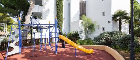 A playground with a slide and climbing structures, surrounded by trees and a white building with a 'M' on it.
