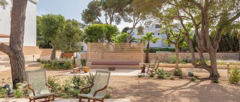 Un área de estar al aire libre con sillas y una mesa, rodeada de árboles y un edificio blanco.