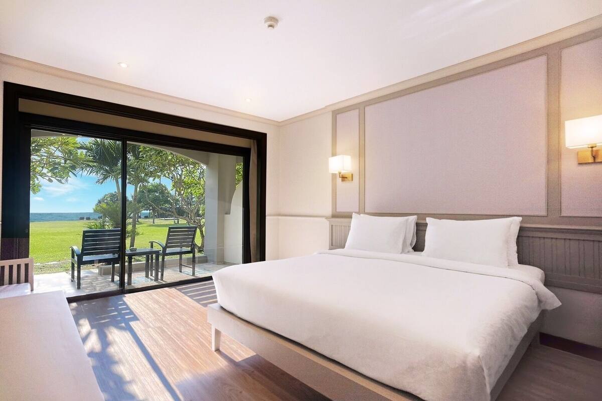 A hotel room with a large bed, a desk, and a sliding glass door leading to an outdoor area with chairs and a view of the ocean.