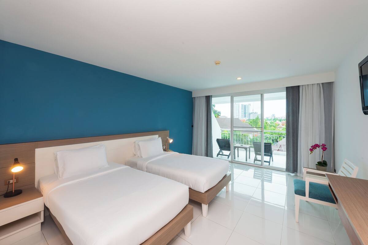 A hotel room with two beds, a desk, and a balcony with chairs.