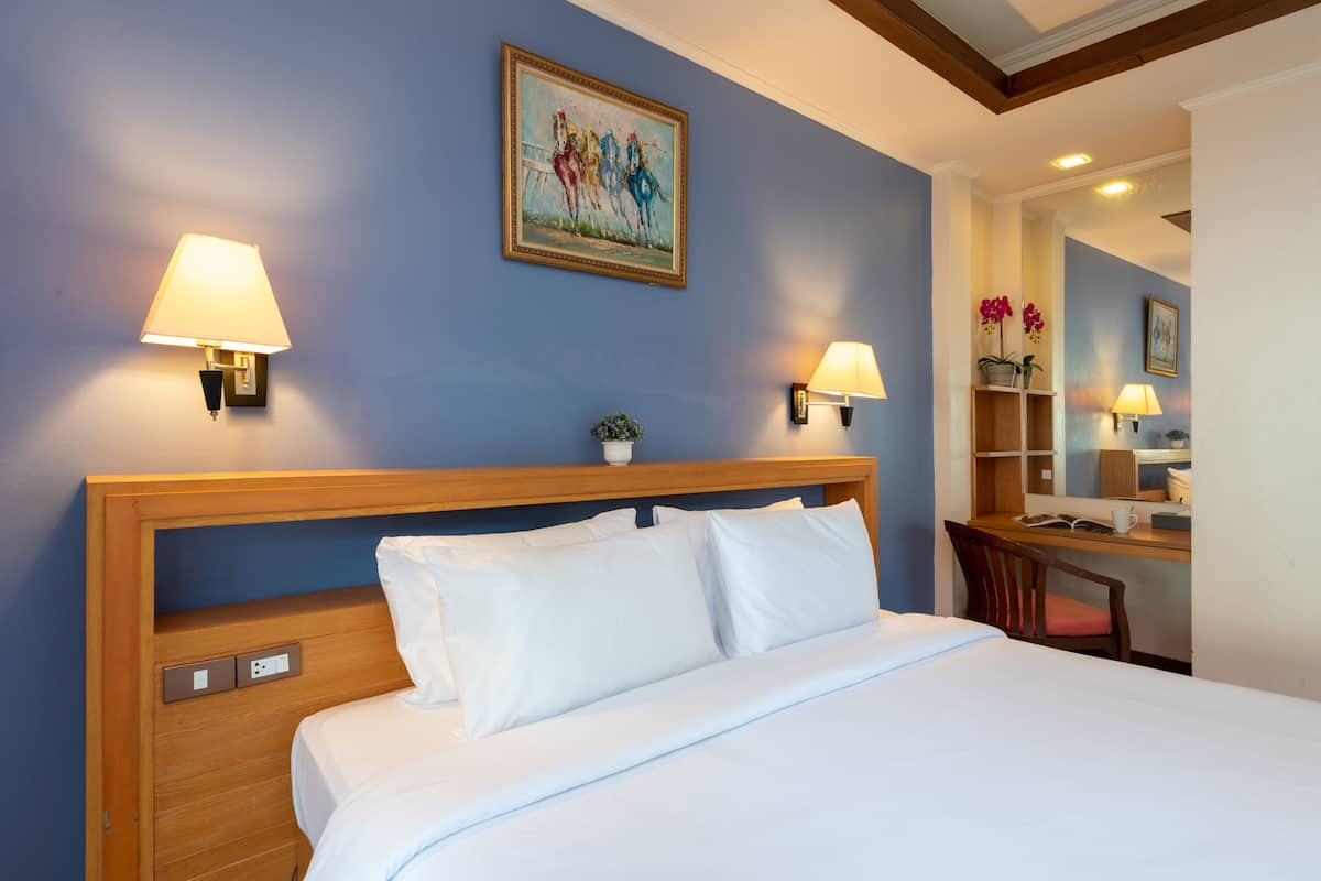 A hotel room with a wooden headboard, a bed with white linens, two wall-mounted lamps, a painting, a desk with a chair, and a small plant.