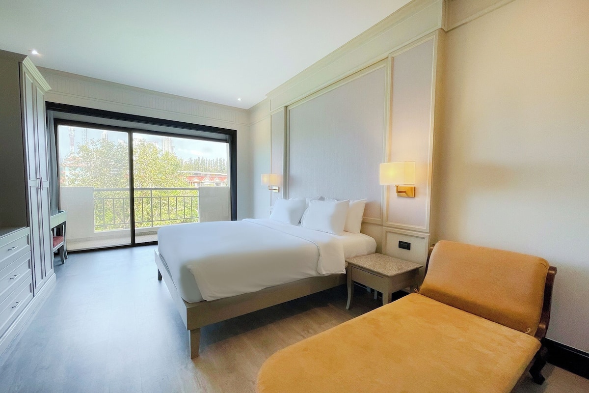 A hotel room with a large bed, a bedside table, a chair, and a view of the outdoors through a sliding door.