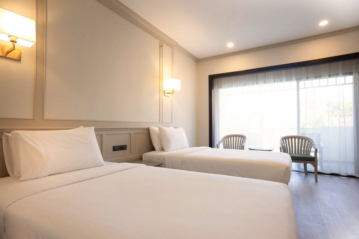 A hotel room with two beds, a small table, two chairs, and a large window with curtains.