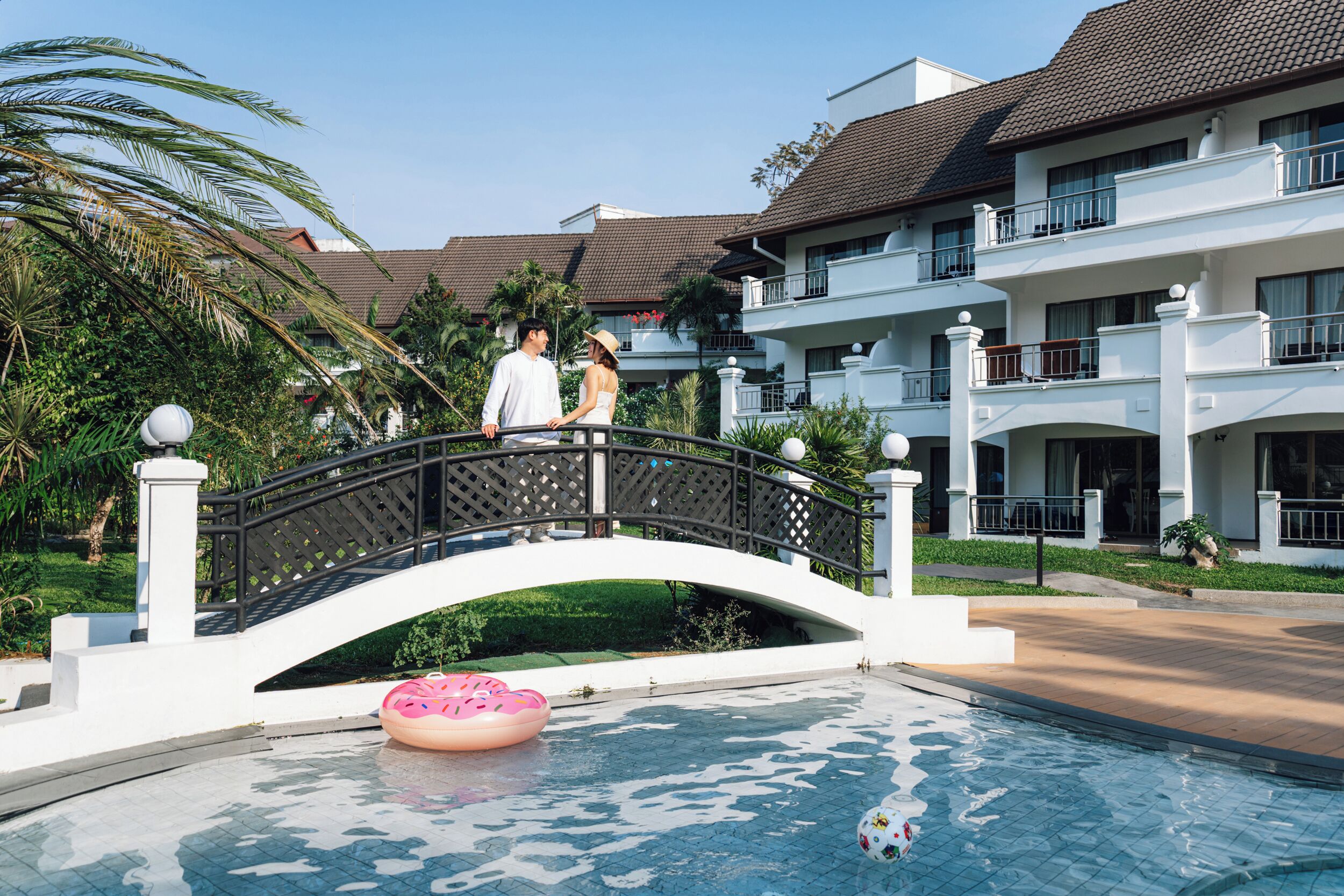 Zona de piscina con un puente, palmeras y un edificio con balcones.