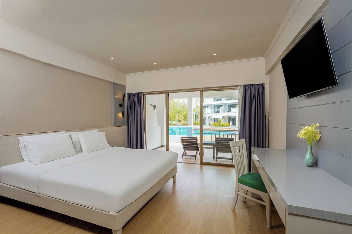 A hotel room with a bed, a desk, a chair, a TV, and a view of a pool and building.