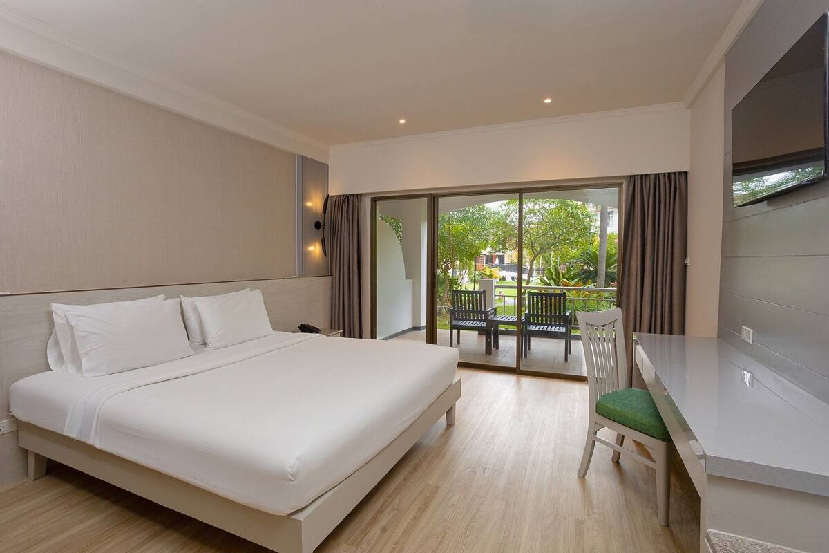 A hotel room with a bed, a desk, and a chair. There is a balcony with a view of trees and buildings.