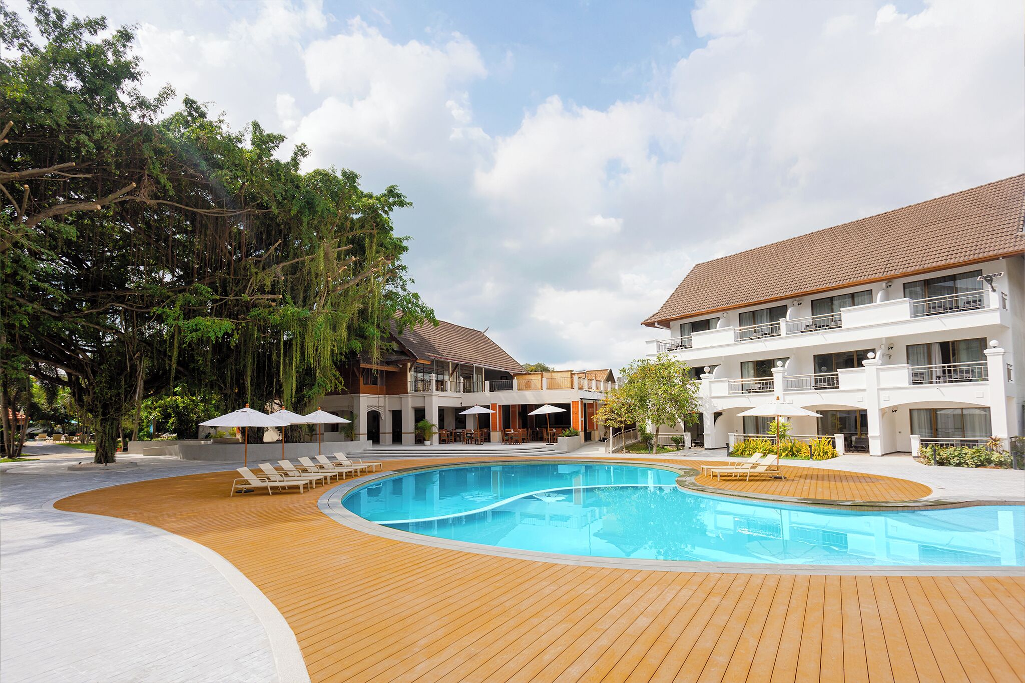A pool area with wooden decking, lounge chairs, and umbrellas, surrounded by trees and a multi-story building with balconies.