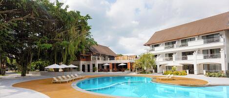 A pool area with wooden decking, lounge chairs, and umbrellas, surrounded by trees and a multi-story building with balconies.