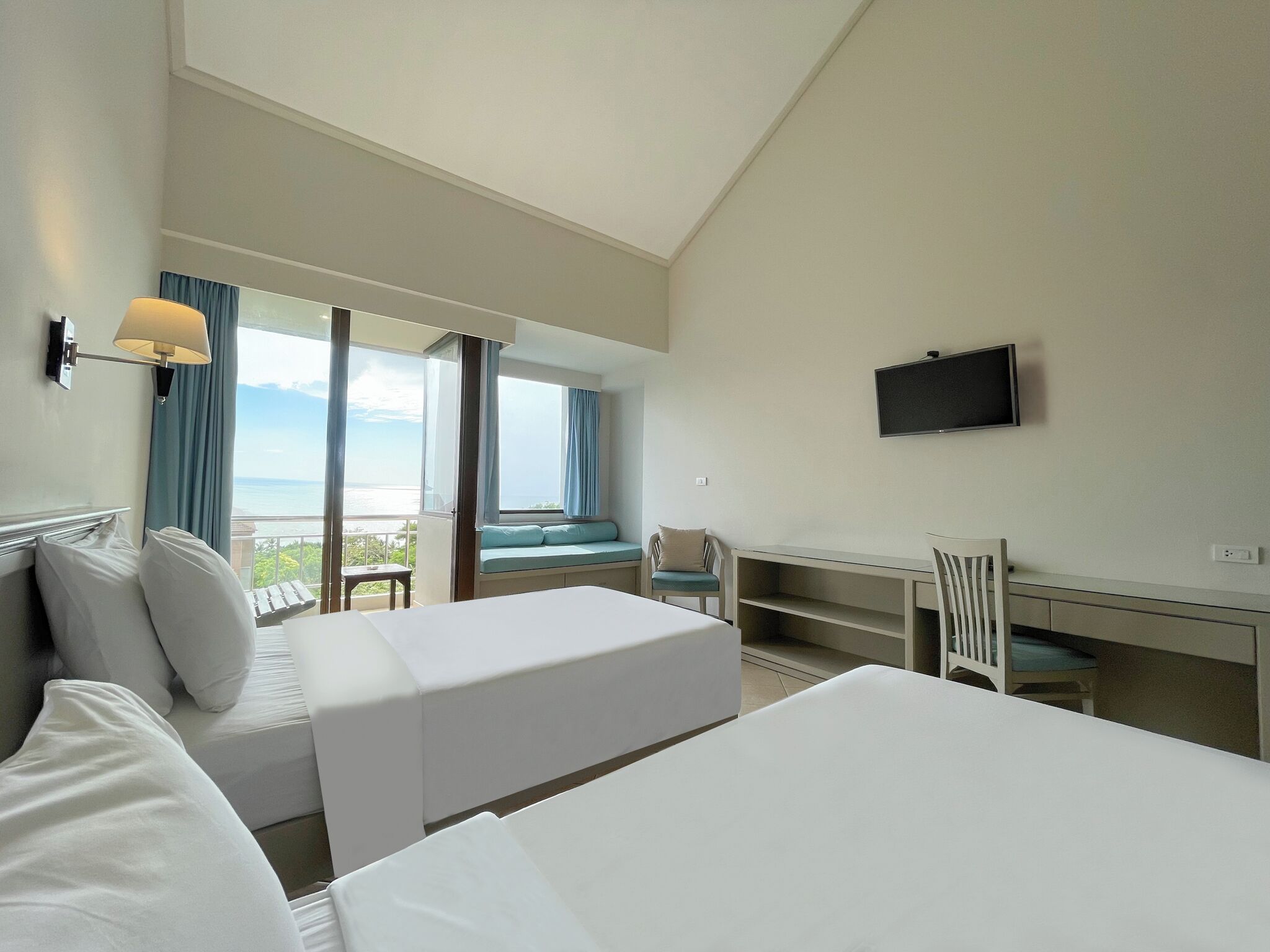 A hotel room with two beds, a desk, a chair, a TV, and a balcony with a view of the sea.
