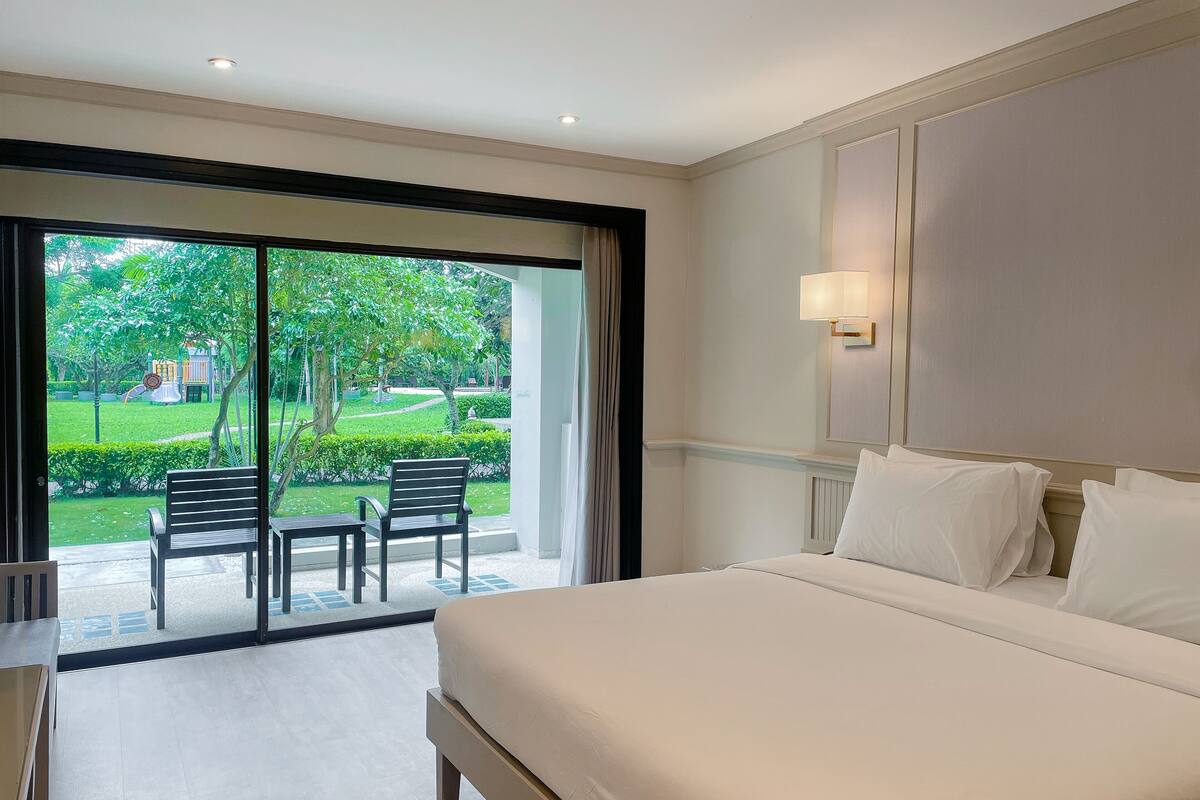 A hotel room with a large bed, a desk, and a chair. There is a view of a garden and a playground outside the window.