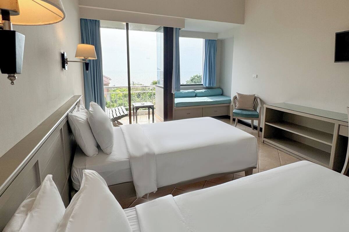 A hotel room with two beds, a desk, a chair, and a view of the sea.
