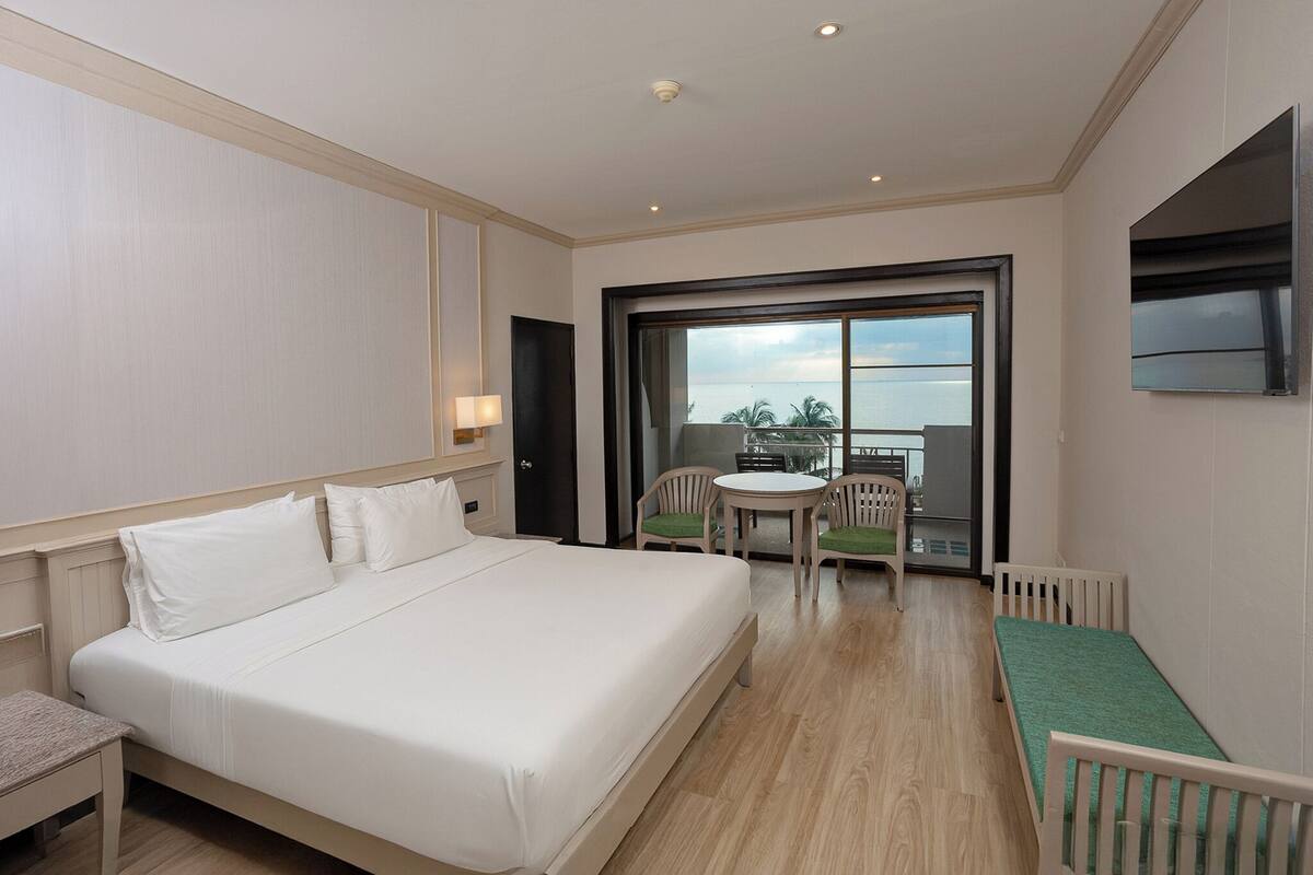 A hotel room with a large bed, a small table with chairs, a balcony with a table and chairs, and a TV mounted on the wall.