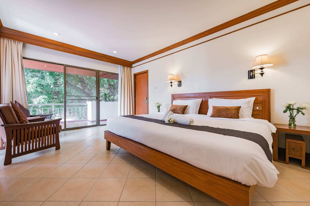 A spacious bedroom with a large bed, wooden headboard, and a bench. There are two wall-mounted lamps and a large window with curtains.
