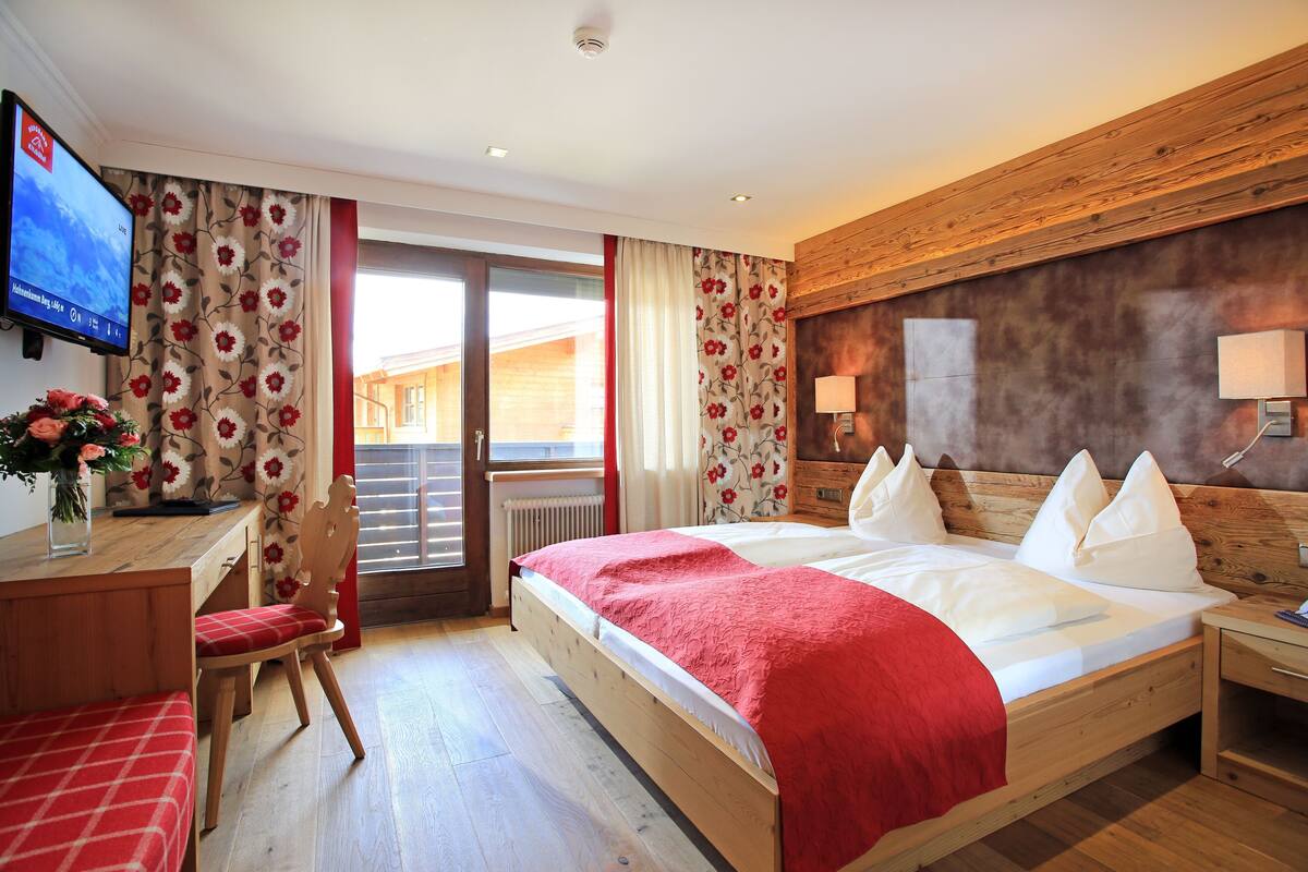 A hotel room with a wooden bed, a desk with a chair, a TV on the wall, and a view of the outside.