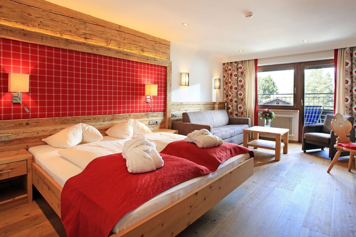 A hotel room with a wooden bed, a sofa, a small table, and a chair. There are two wall-mounted lamps and a window with curtains.
