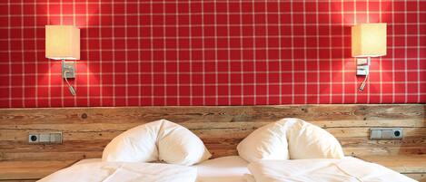 A bed with white linens and a red blanket, a wooden headboard, and a red plaid wall with two wall-mounted lamps.