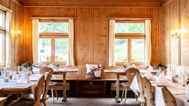 Un interior de madera con mesas dispuestas para comer, ventanas amplias y lámparas fijadas a la pared.