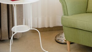 Una habitación con un sillón verde, una lámpara blanca y un jarrón de vidrio con tulipanes naranjas sobre una pequeña mesa blanca.