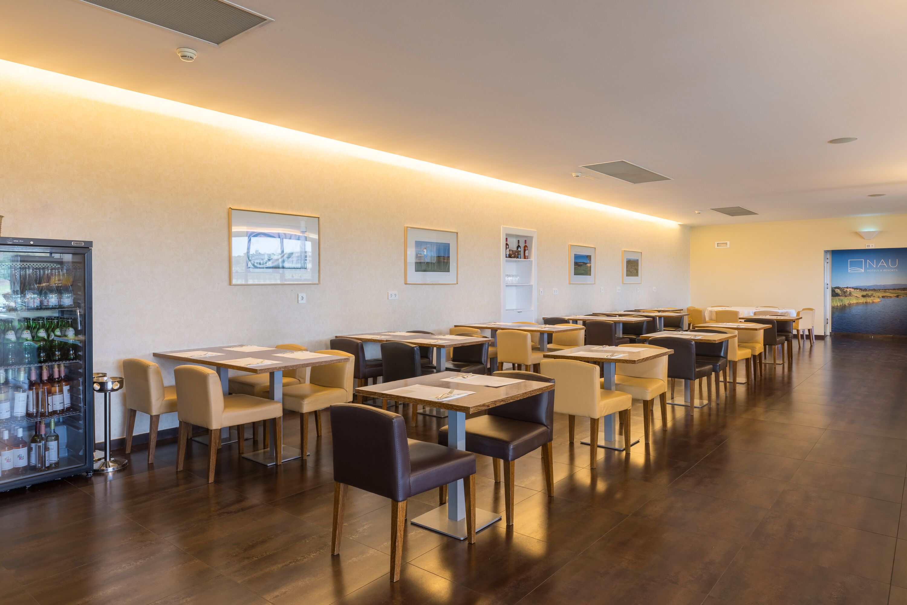 A restaurant with multiple tables and chairs, a refrigerator with bottles, and framed pictures on the wall.