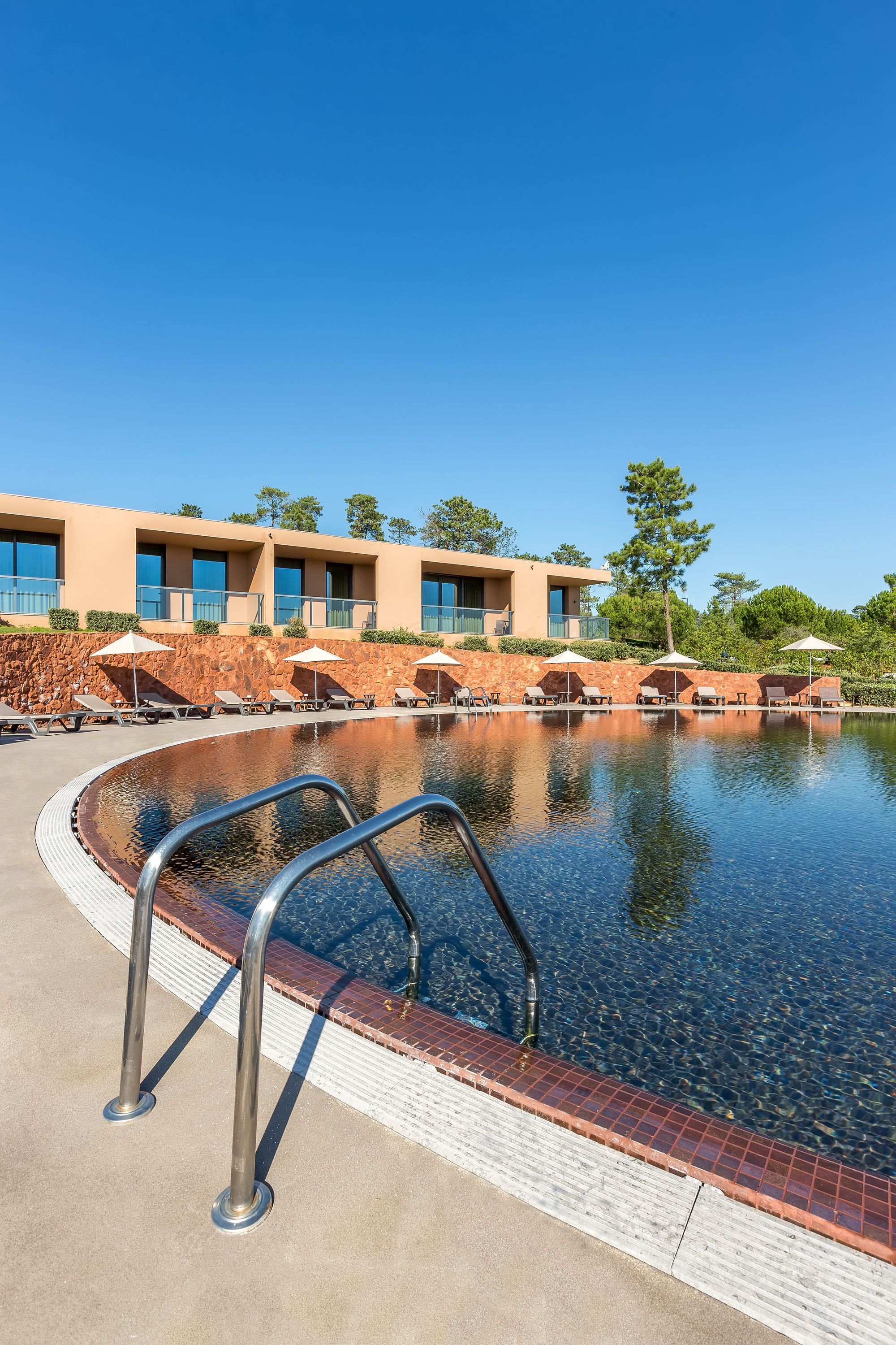 Un área de piscina con sillones de descanso y sombrillas, un muro de piedra y un edificio con varias ventanas y balcones.