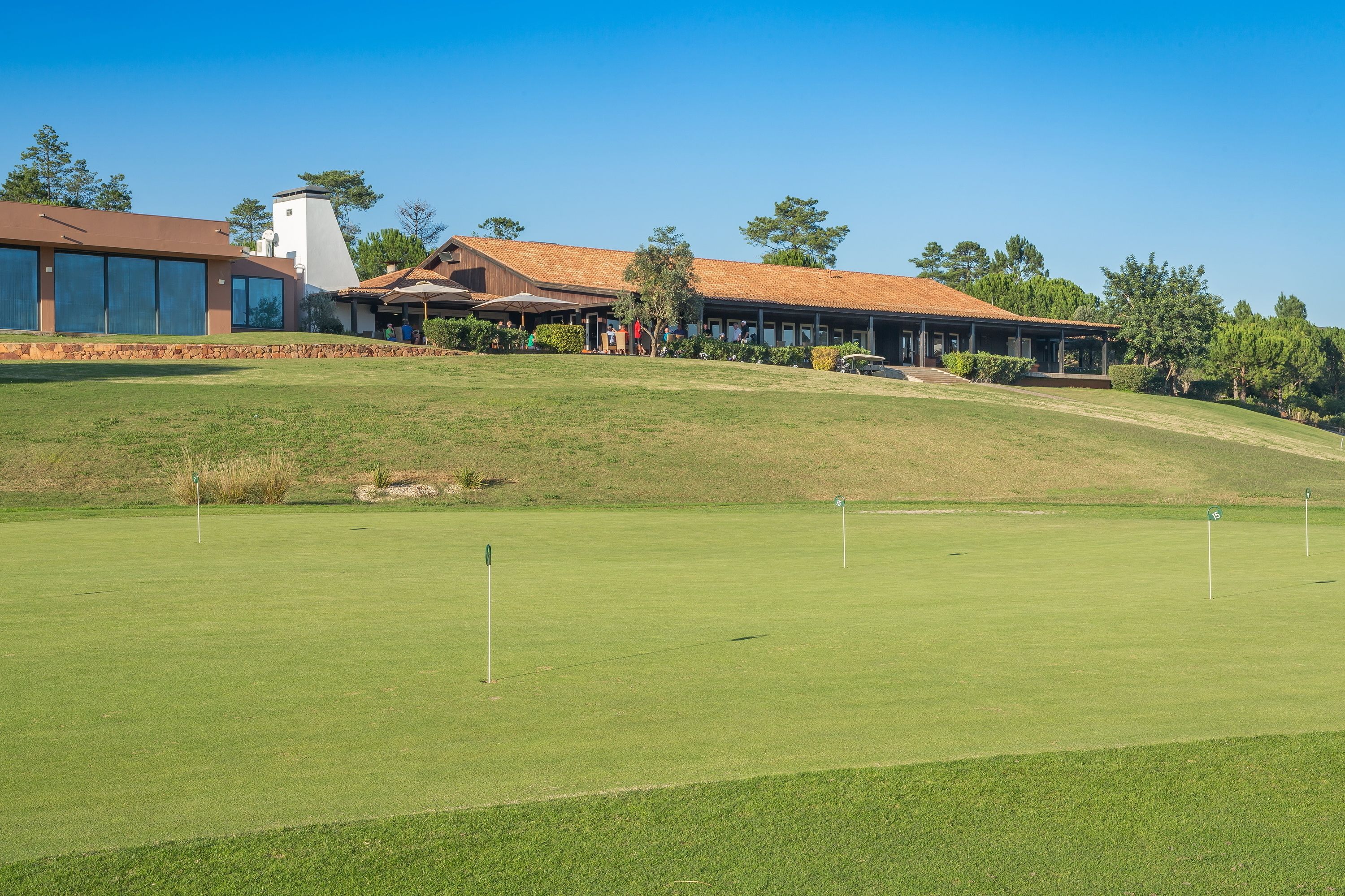 Un parcours de golf doté d’un club-house, d’un grand bâtiment à toit en pente et d’une colline plantée d’arbres.