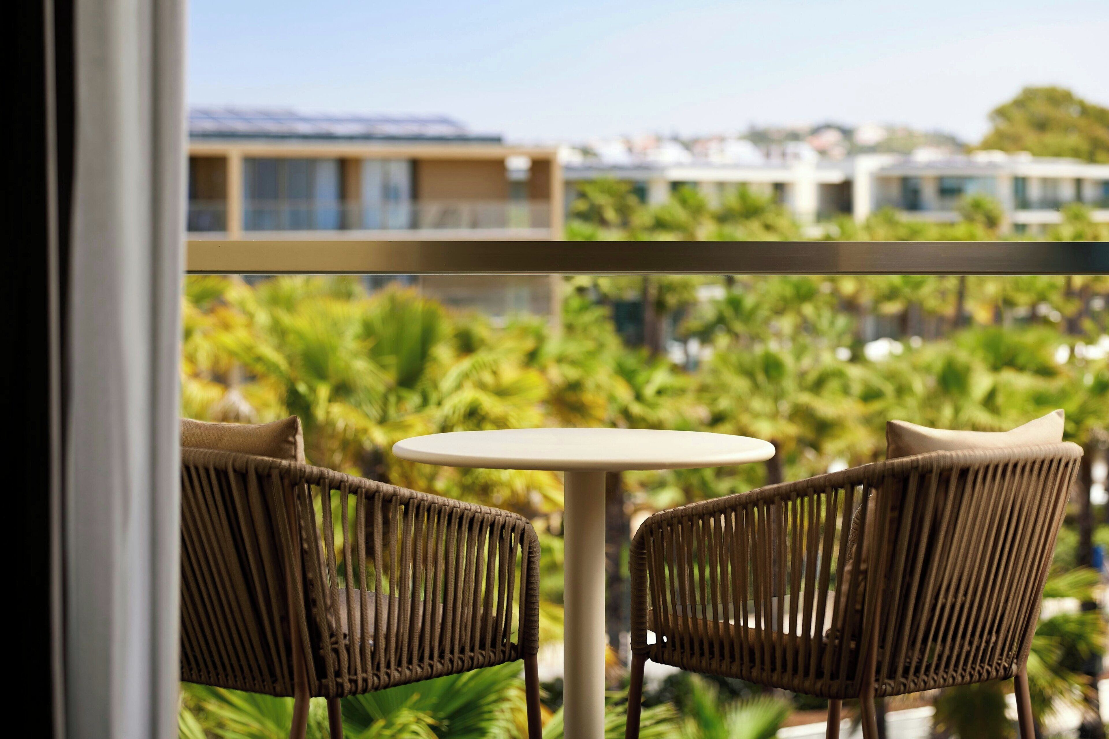 A balcony with wicker chairs and a small round table, overlooking a lush garden and buildings in the background.