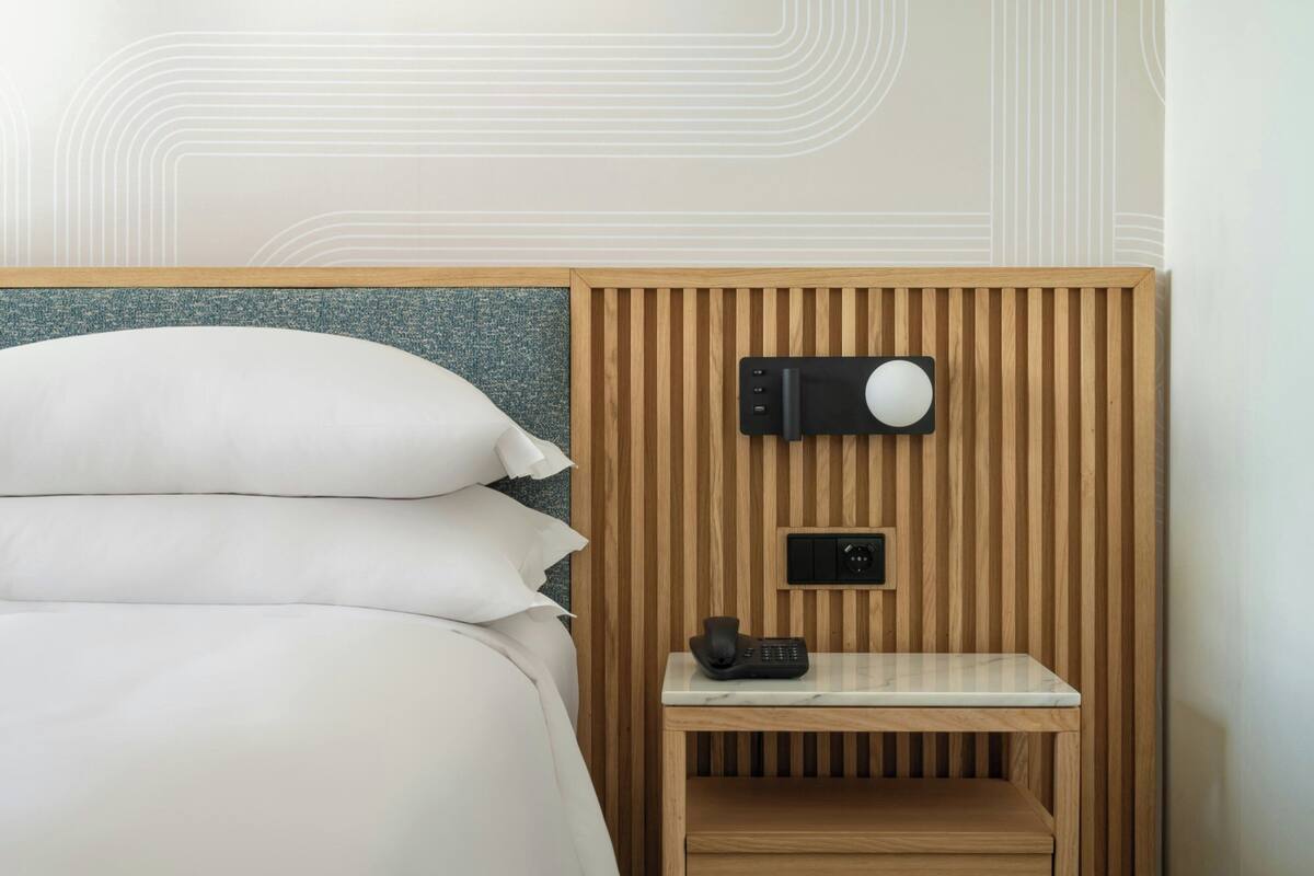 A hotel room with a bed, a bedside table with a phone, and a wall-mounted speaker.