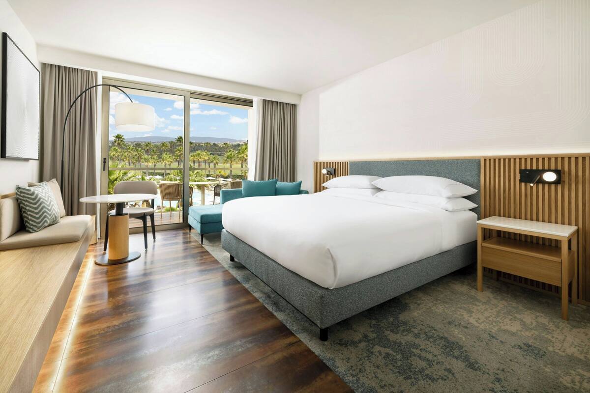 Habitación de hotel moderna con una cama grande, un sofá, una mesita y vistas a palmeras y montañas a través del ventanal.