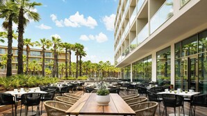 An outdoor dining area with multiple tables and chairs, surrounded by palm trees and a modern building with large windows. - Algarve Marriott Salgados Golf Resort & Spa (Albufeira)