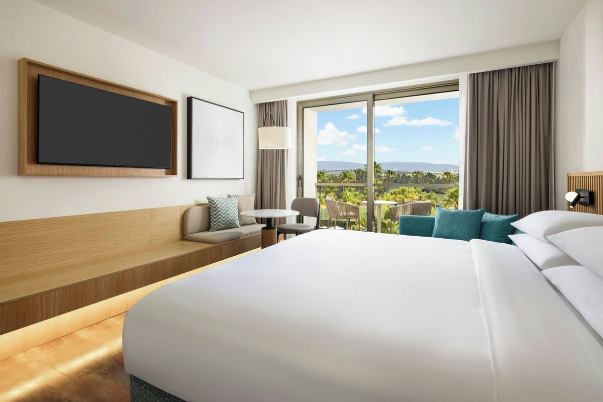 Habitación de hotel moderna con una cama grande, un sofá, una mesita y una silla. Una ventana grande ofrece vistas al exterior.