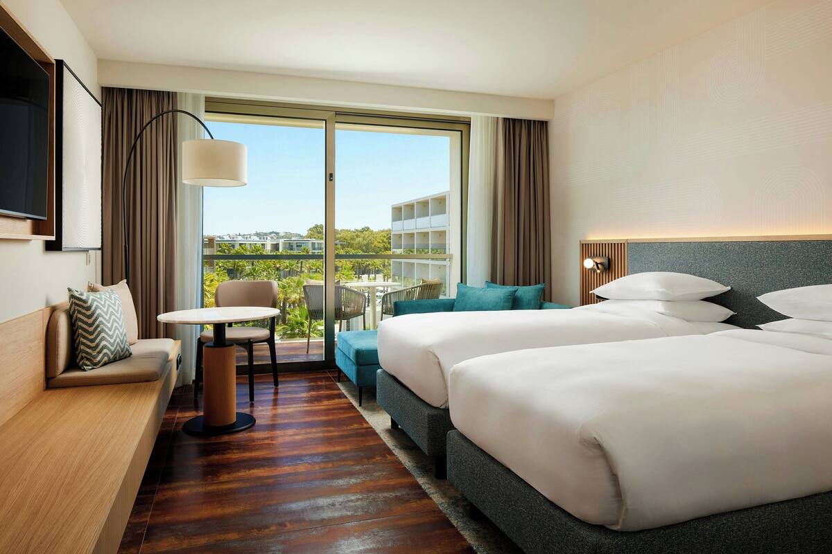 Habitación de hotel con dos camas, un sofá, una mesita y un balcón con vistas a edificios y vegetación.