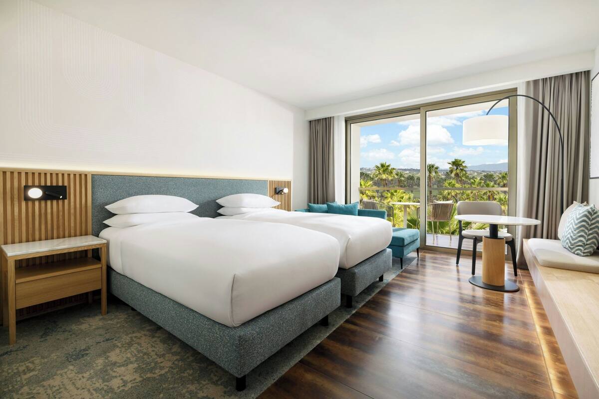 Habitación de hotel con una cama grande, un sofá, una mesita y vistas a palmeras.