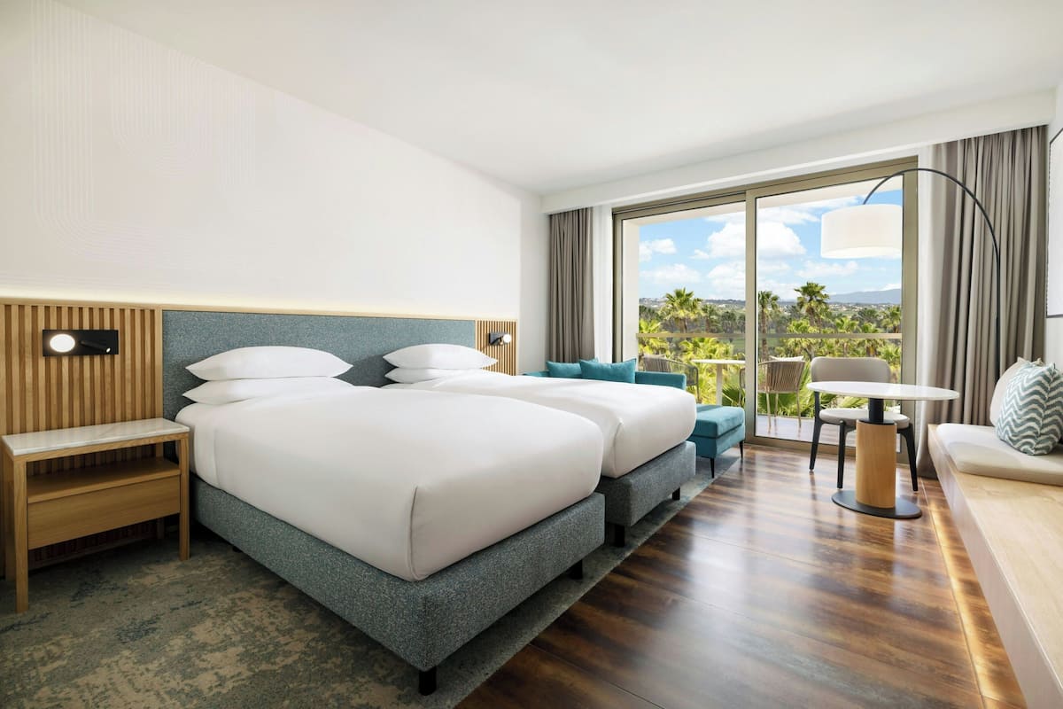 Ein Hotelzimmer mit einem großen Bett, einer Sitzecke, einem kleinen Tisch und Blick auf Palmen.