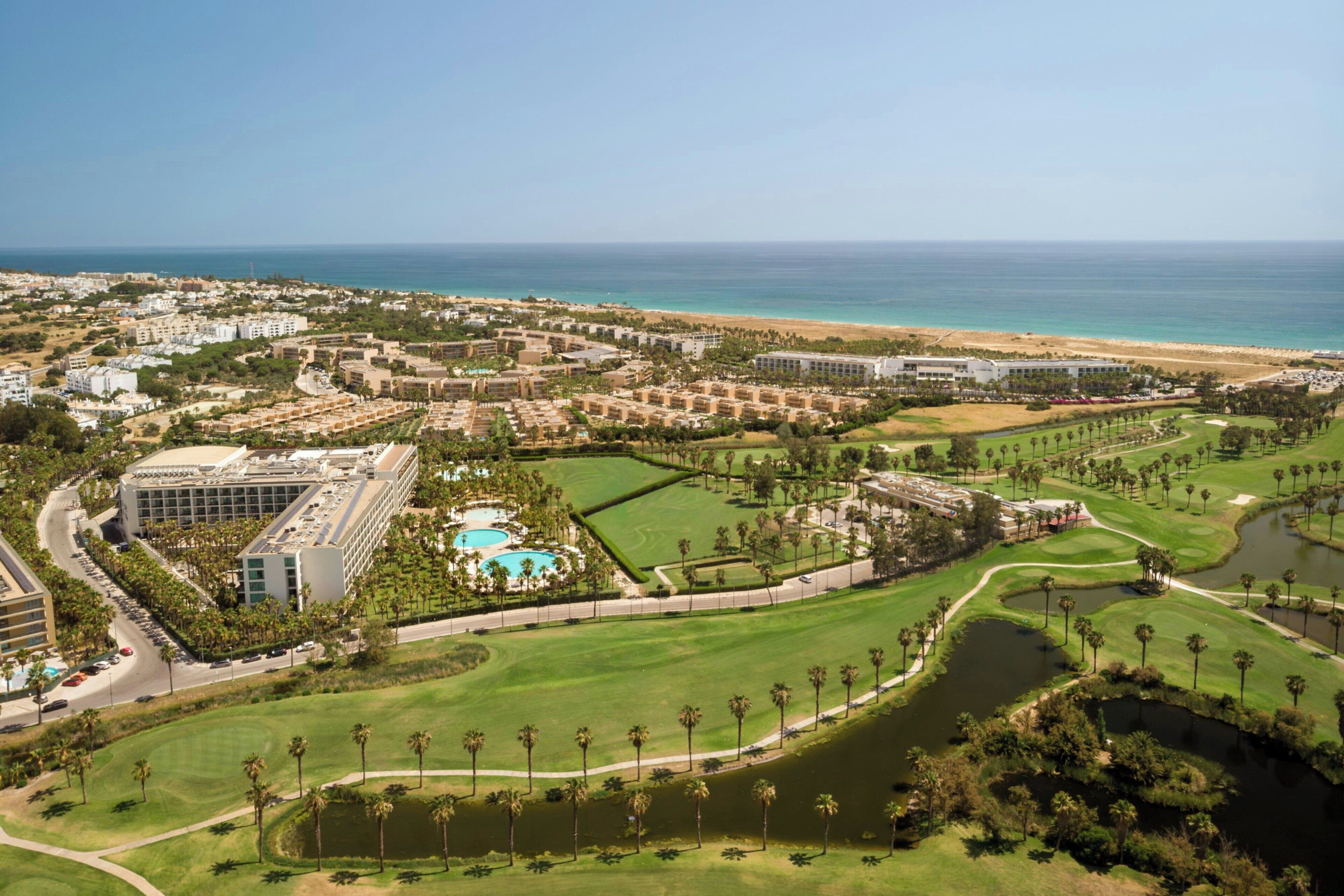 A golf course with a large building complex and a beach in the background.
