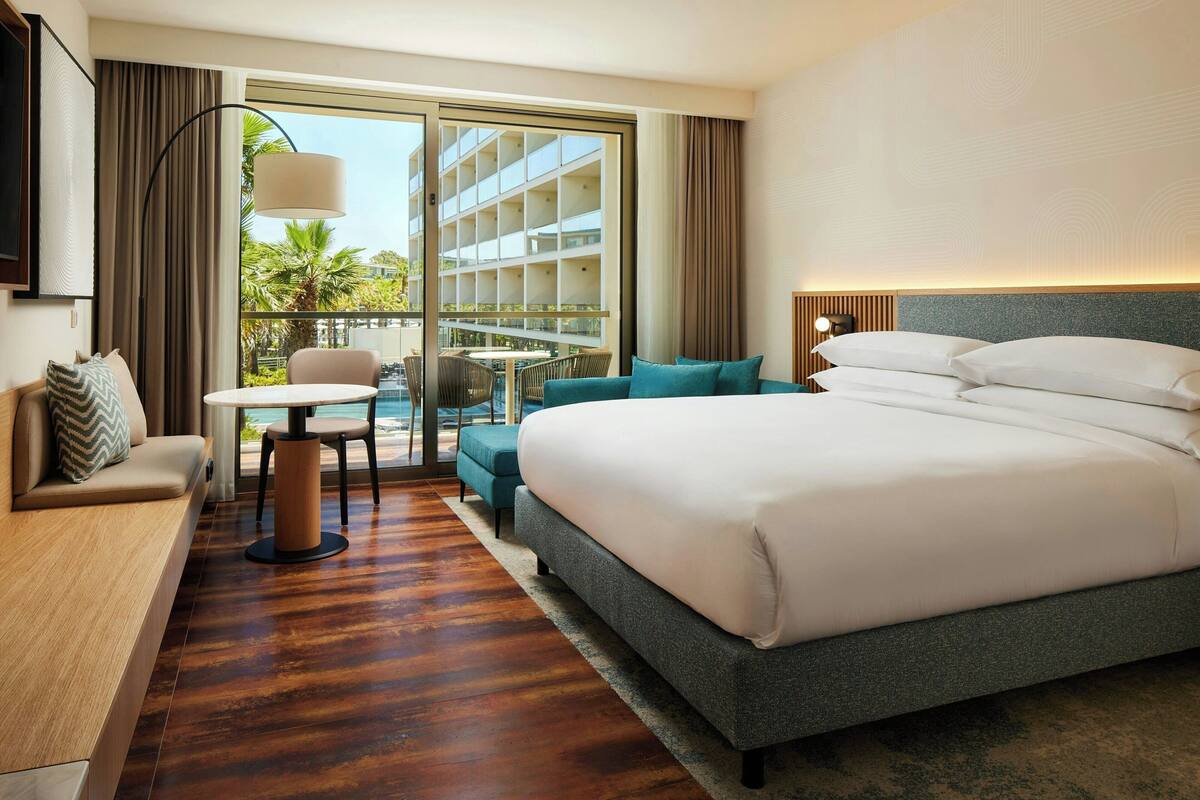 Habitación de hotel con una cama grande, un sofá, una mesita y un balcón con vistas a palmeras y un edificio.