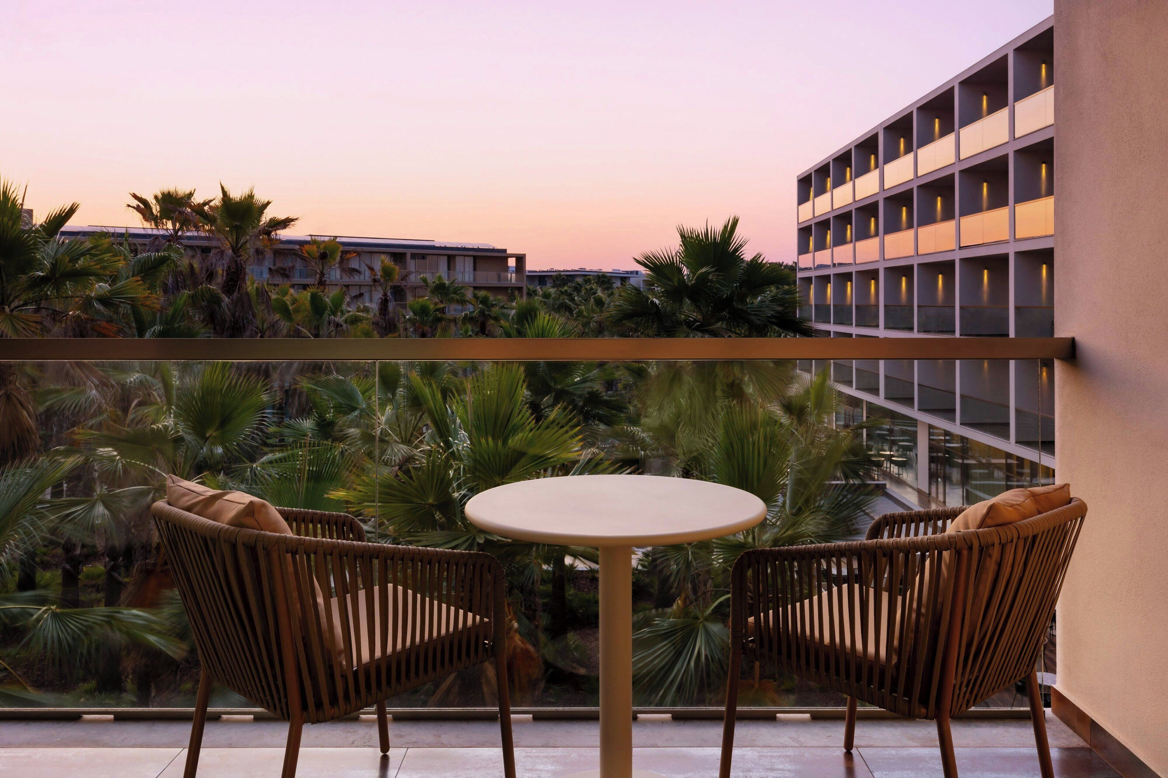 A balcony with two wicker chairs and a small round table, overlooking a building and palm trees.