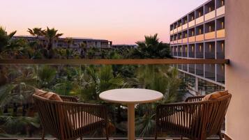 A balcony with two wicker chairs and a small round table, overlooking a building and palm trees.