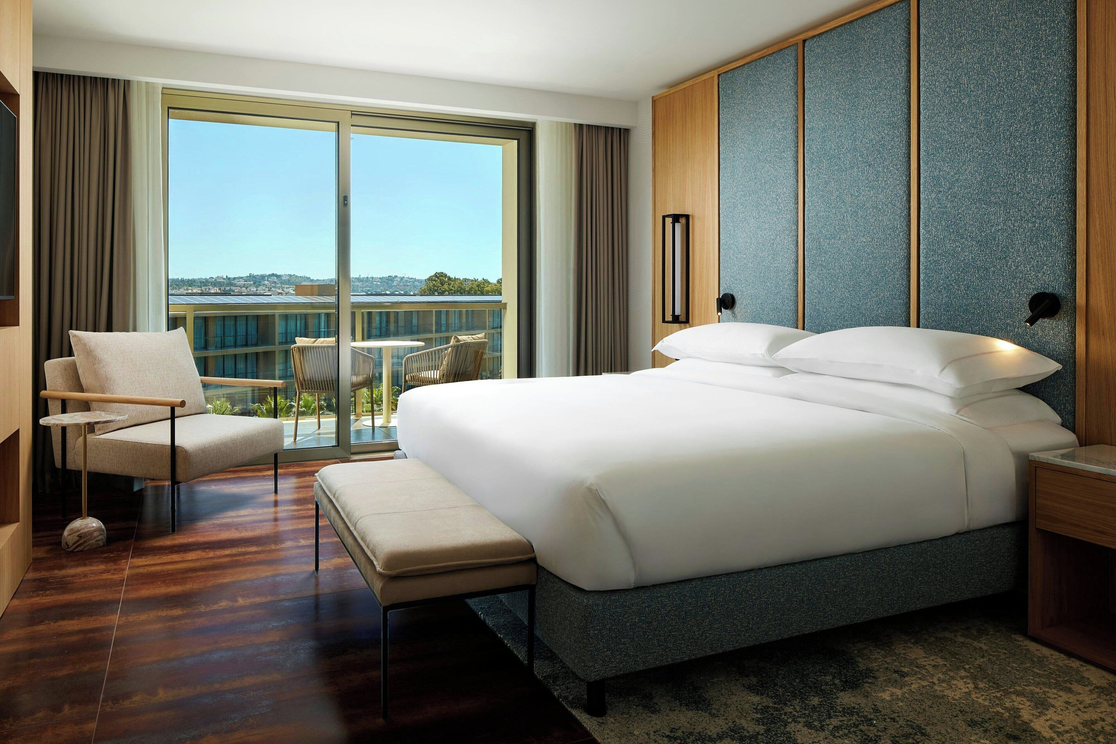 A modern hotel room with a large bed, a chair, a small table, and a view of the city through large windows.
