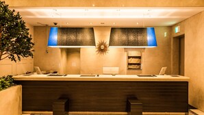 A reception desk with a modern design, a large decorative sunburst mirror, and a potted plant to the left.