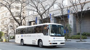 A white bus with "HOTEL MYSTAYS PREMIER Omori" written on the side, parked on a street with buildings and trees in the background.
