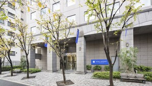A hotel entrance with a sign, paved walkway, benches, and trees.