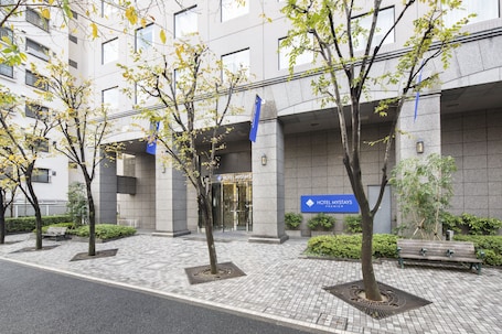 A hotel entrance with a sign, paved walkway, benches, and trees.