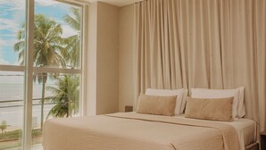 Habitación de hotel con un ventanal grande, cama con ropa de cama color crema y vista a palmeras y al océano.