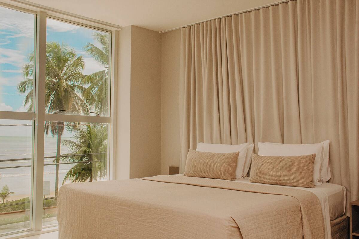 A hotel room with a large window, a bed with beige bedding, and a view of palm trees and the ocean.