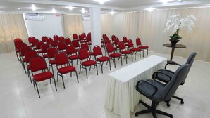 Un salón de conferencias con filas de sillas rojas, una mesa blanca y una orquídea en maceta.