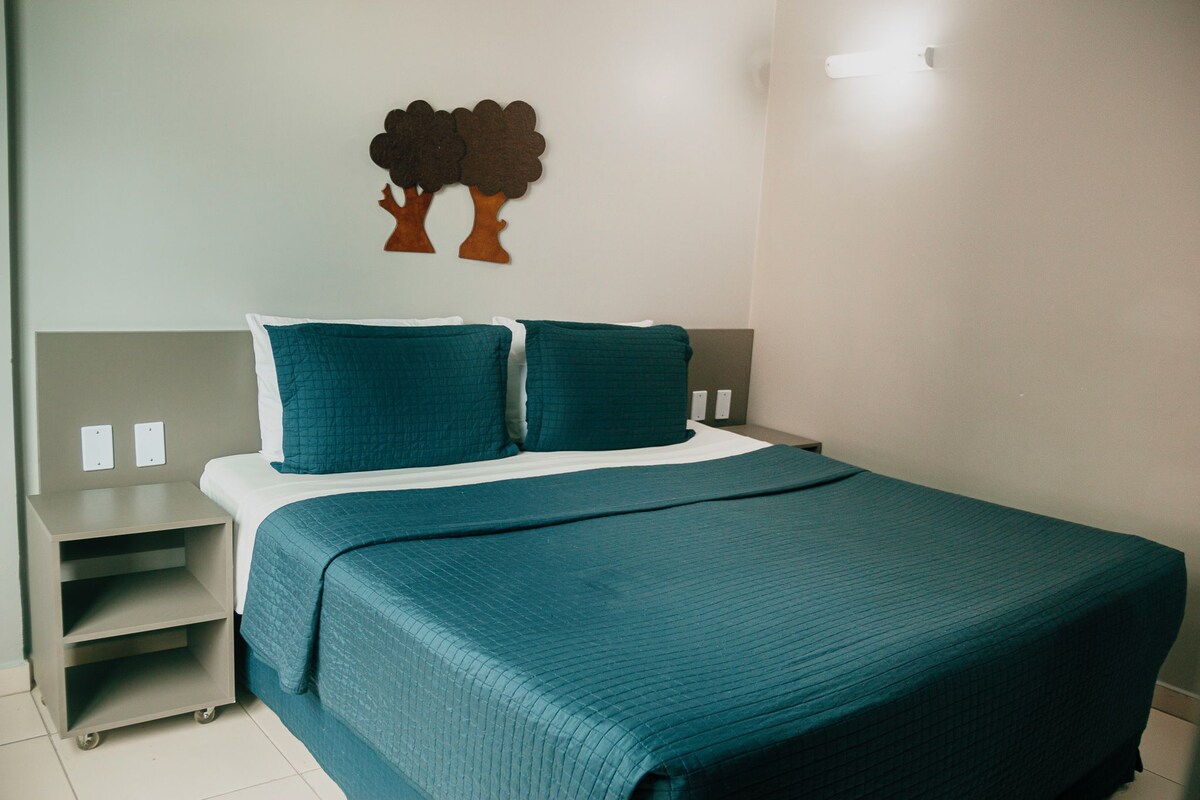 A neatly made bed with a teal bedspread and pillows, a small bedside table, and a wall-mounted tree decoration.