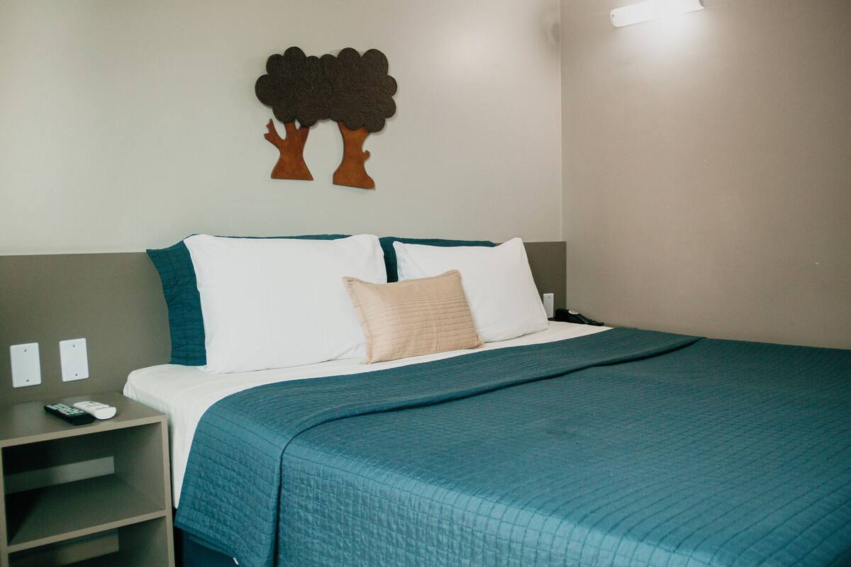 A neatly made bed with a teal bedspread and white pillows, a small bedside table with a remote control, and a wall-mounted tree decoration.